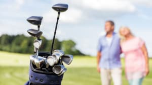 A set of golf clubs in a bag is in focus in the foreground, while an older man and woman walk together on a golf course, blurred in the background.